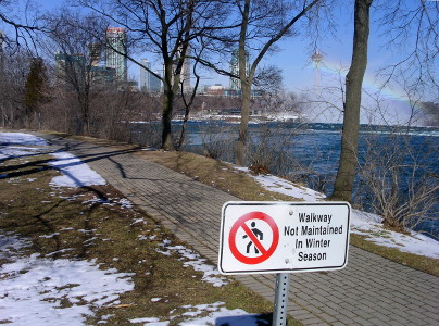 [Just behind the sign in the image is a brick-paved walkway on a hillside above the water leading to Horseshoe Falls. There is quite a bit of brown grass visible amid the snow on the grass, but the walkway is completely clear of snow.]